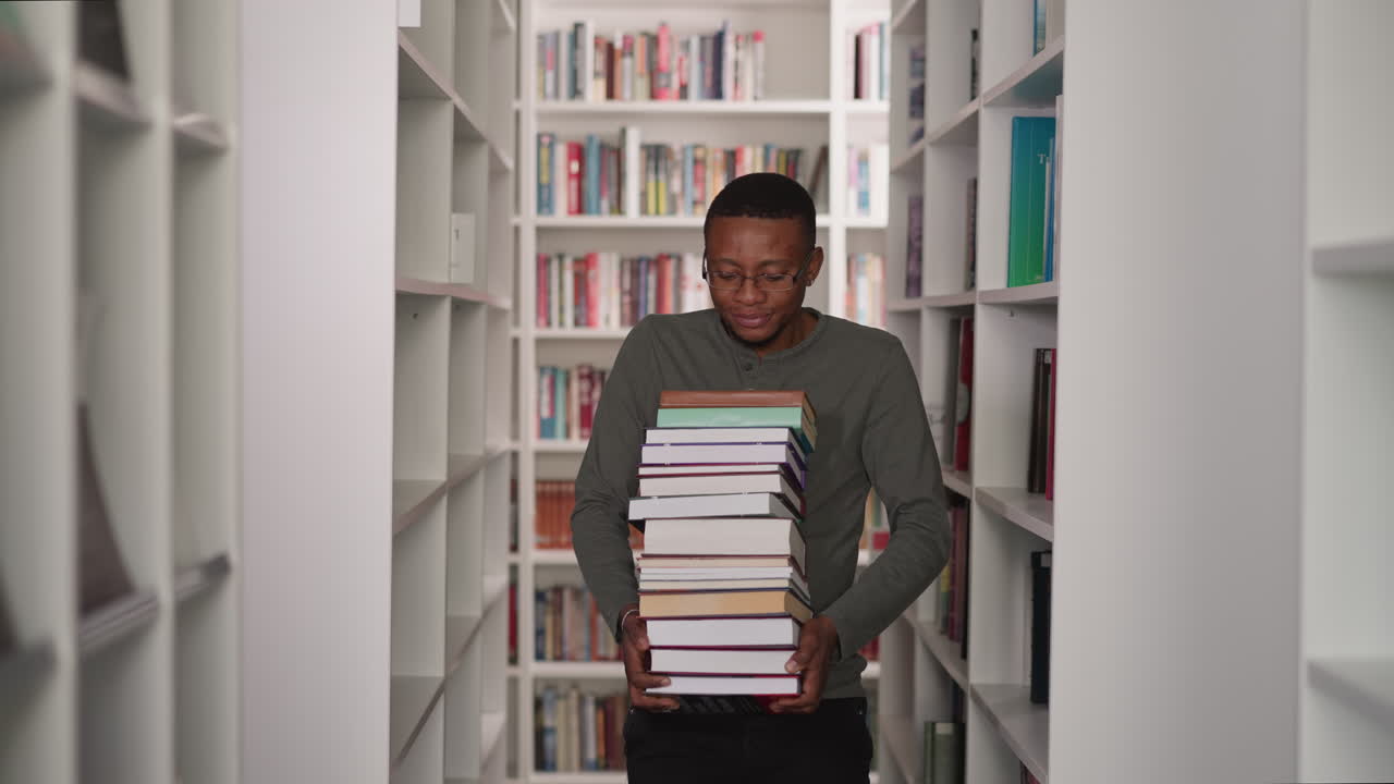 Guy carries heavy book stack in library. African American student walks with large textbooks pile along aisle between bookshelves. Information store