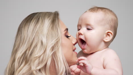 mother adoring her baby boy kisses him on the cheek. cute little infant smiling from mom's kisses. close up. white backdrop.
