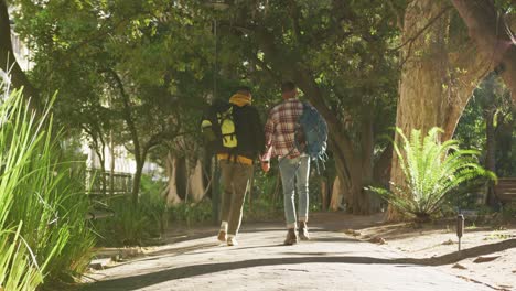 Back-view-of-two-happy-mixed-race-gay-male-couple-walking-and-talking-in-park-with-backpacks