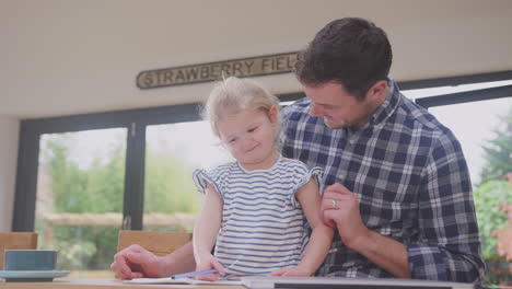 father at home at kitchen counter helping young daughter to draw picture in book - shot in slow motion