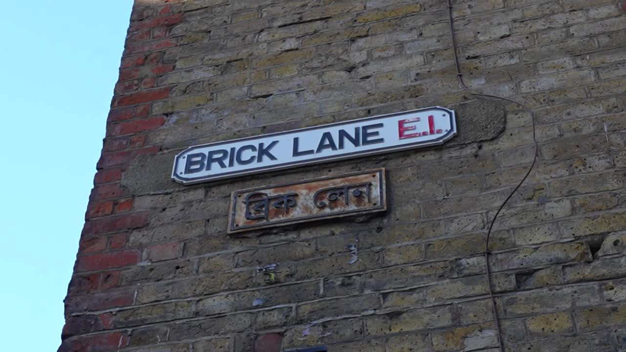 Brick Lane street sign on a brick wall with Bangladeshi nameplate below under clear sky