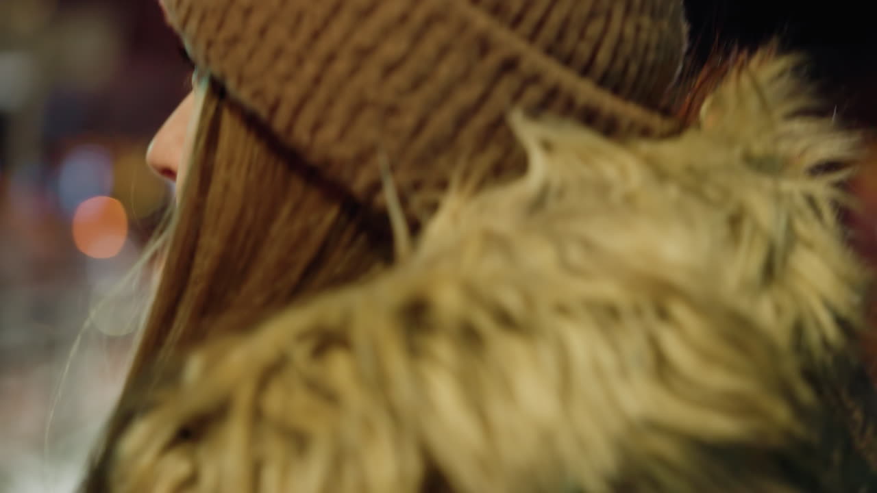 вид от первого лица женщины в зимнем пальто с меховым капюшоном и коричневым обогревателем головы, пересекающей дорогу ночью. в фоновом режиме видны размытые городские огни и машины