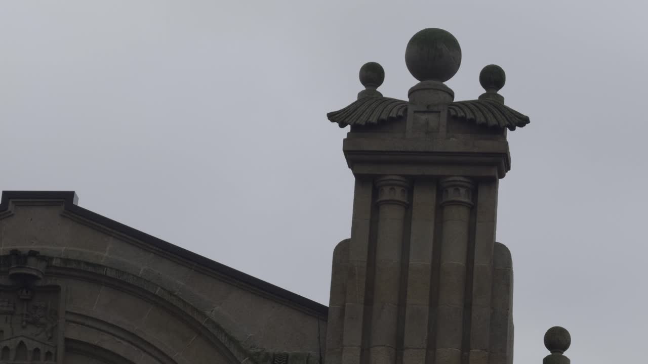 Colonna di pietra con dettagli alati e sfere in cima. Cielo grigio nuvoloso.