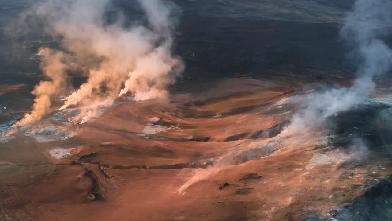 Aerial drone footage showing smoking pillars that are created from hot springs in Iceland or Greenland mountains that have red, orange and black ground during a sunny day. Smoke is white and fluffy.