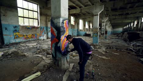 graffiti painter is protective mask and gloves is drawing on old column in dirty empty building using aerosol paint. young man is wearing casual clothes and cap.