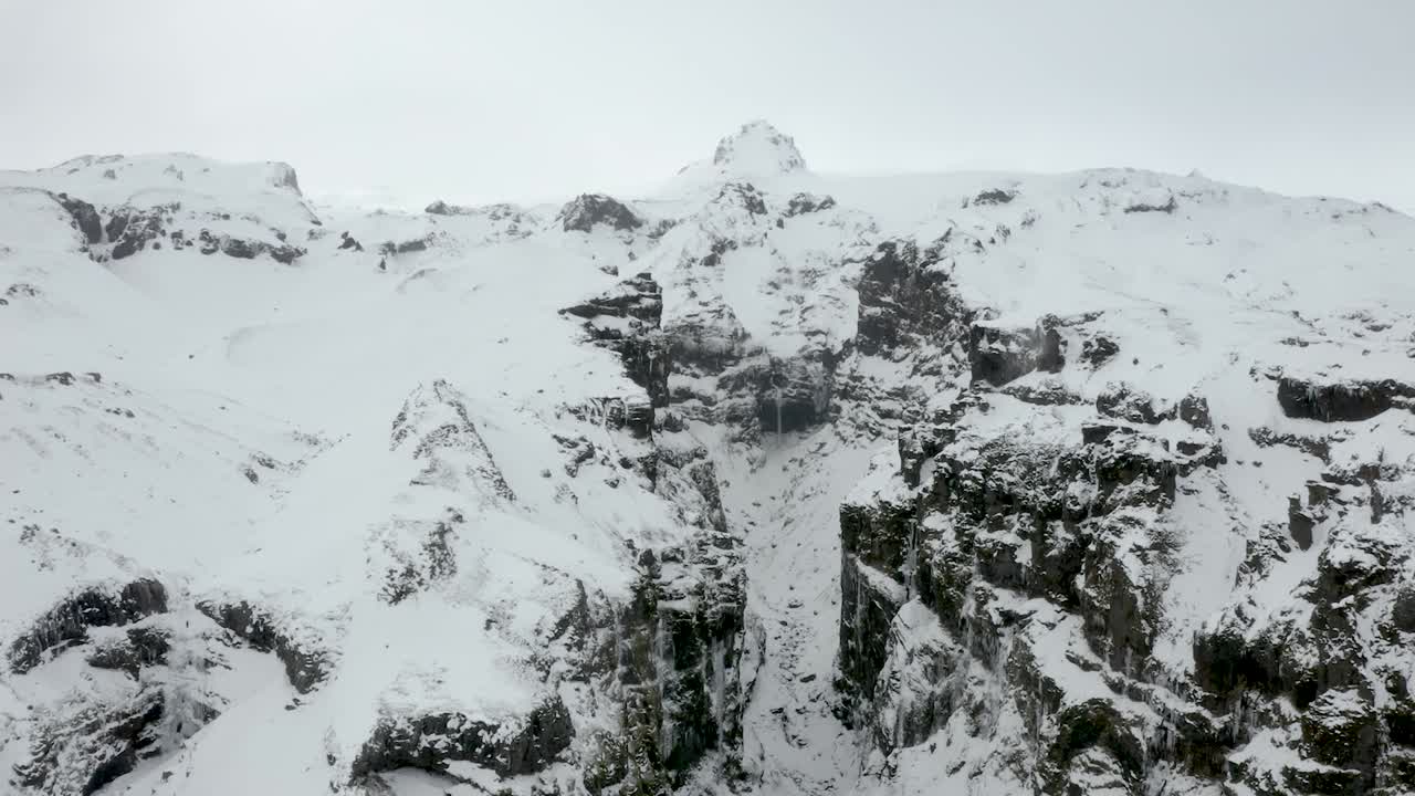 Aerial close up approaching deep frozen snow covered canyon with high rocky cliffs in Iceland.