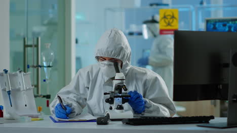 biologist in ppe suit working on microscope and writing information on clipboard