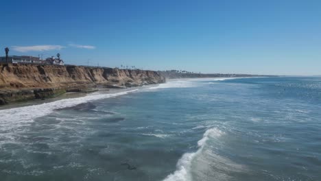 Drone-shot-panning-over-bright-sunny-ocean-coastline-with-beautiful-blue-waves-and-and-a-panorama-of-seaside-cliffs