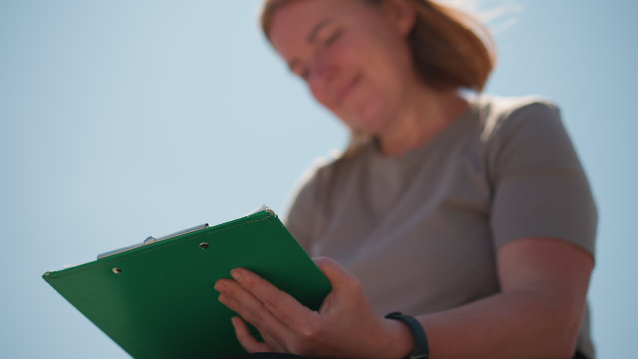 Woman with windblown hair writes on clipboard under bright sky, sunlight reflecting off green cover, face softly blurred in gentle focus, evoking calm concentration