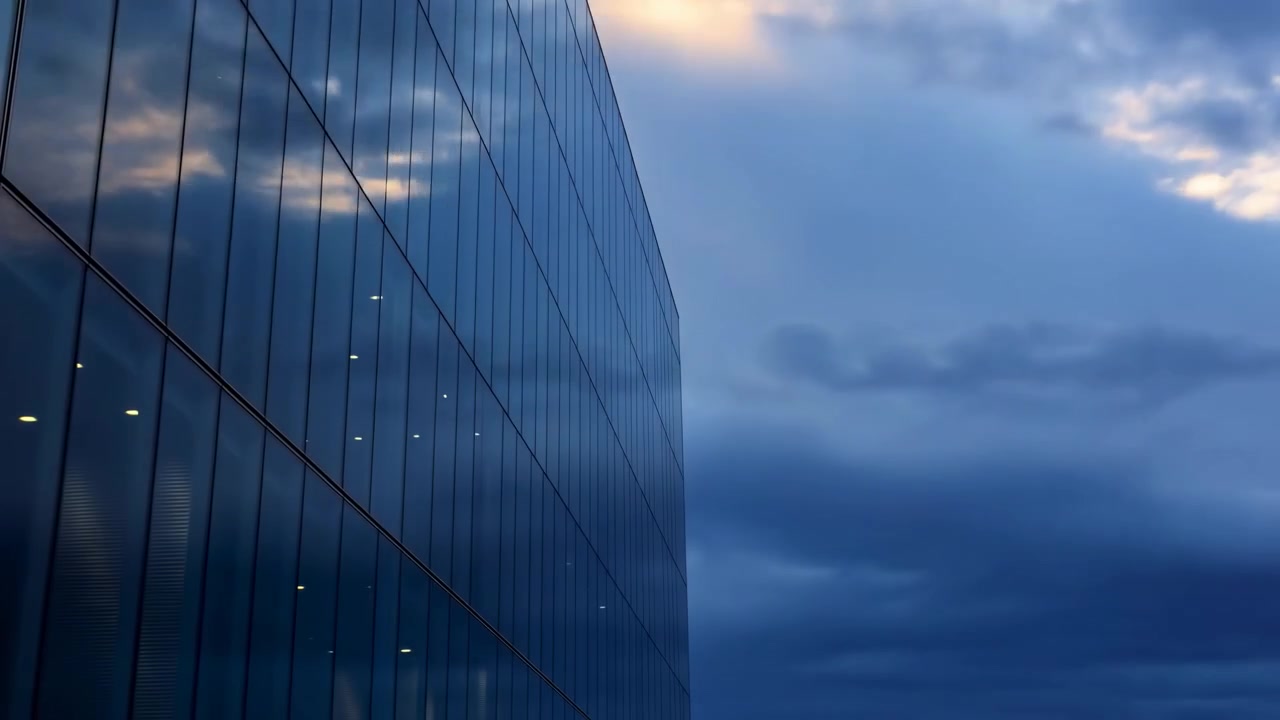 Quadro intermediário mostra uma fachada elegante de vidro de um edifício angulada para a esquerda, captando a luz quente do pôr do sol.