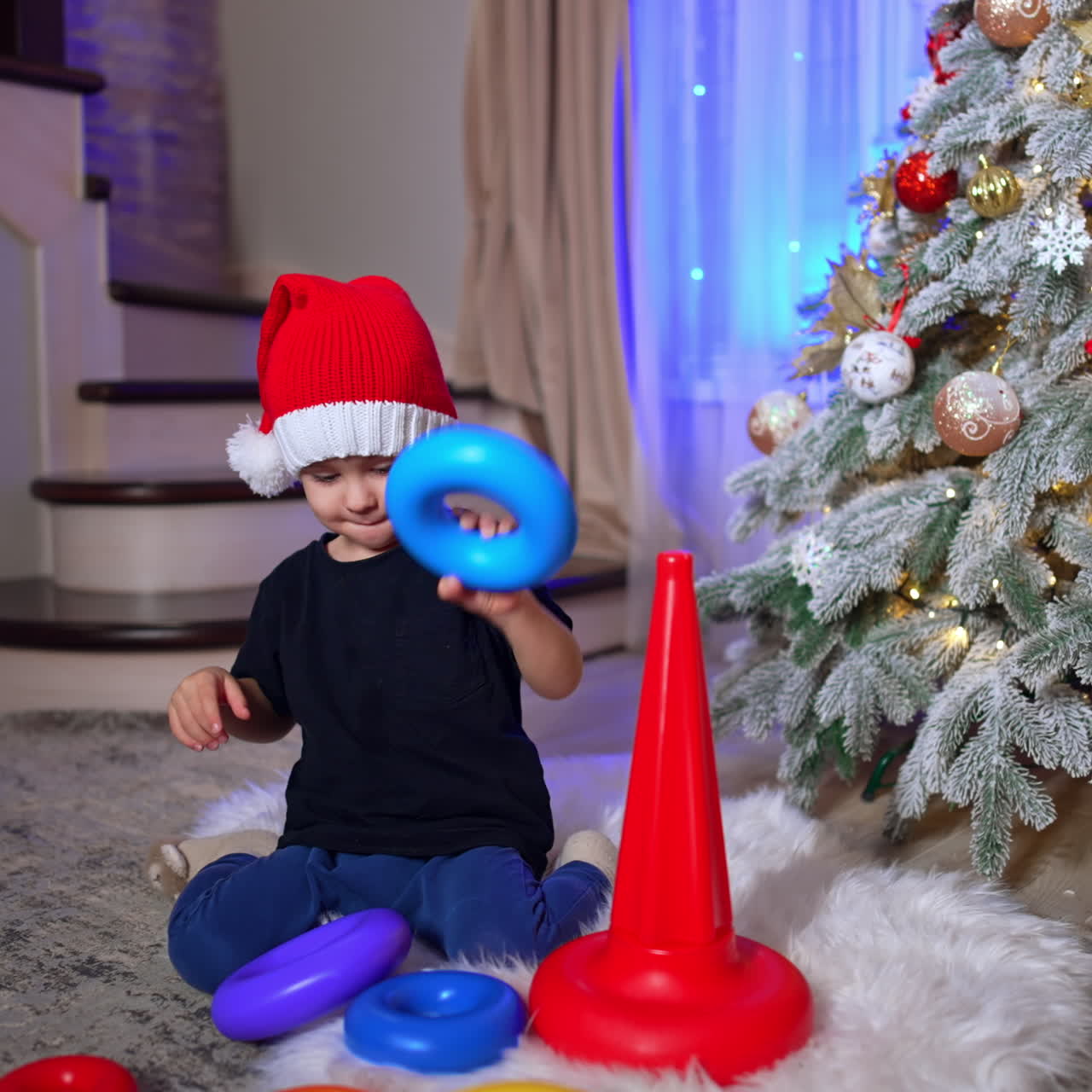 Smart Caucasian toddler wearing Santa cap assembling pyramid. Black cat passes near the kid playing at Christmas tree.