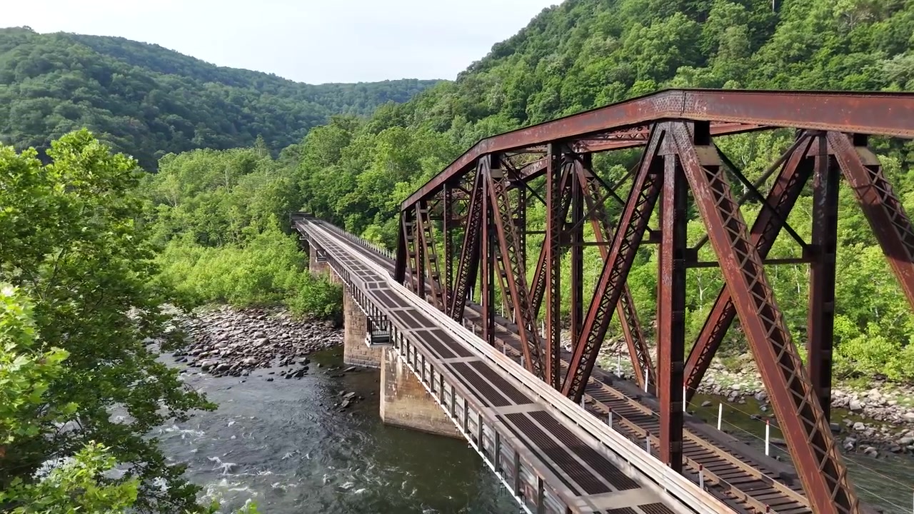Aerial drone perspective of an old steel railway truss bridge crossing the river at New River Gorge National Park, West Virginia. Historic infrastructure surrounded by summer forest.