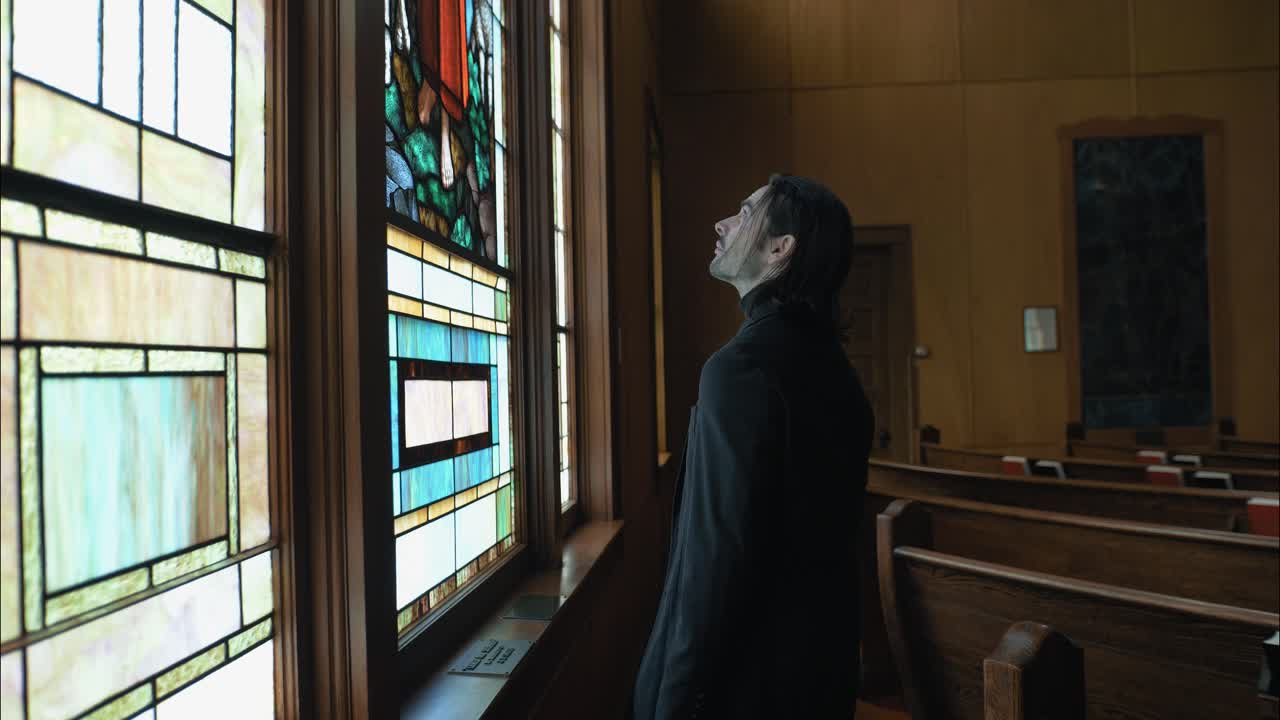 A man stands inside a church, gazing at a stained-glass window of Jesus the Good Shepherd, evoking themes of spirituality, faith, and quiet reflection
