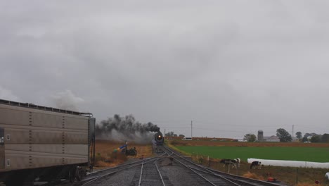 Fahren-Sie-Mit-Blick-Auf-Eine-Dampflokomotive,-Die-Fracht-Zieht-Und-An-Einem-Regnerischen-Tag-Mit-Vieh-Beobachtet,-Das-Auch-Mit-Rauch-Und-Dampf-In-Den-Hof-Zieht