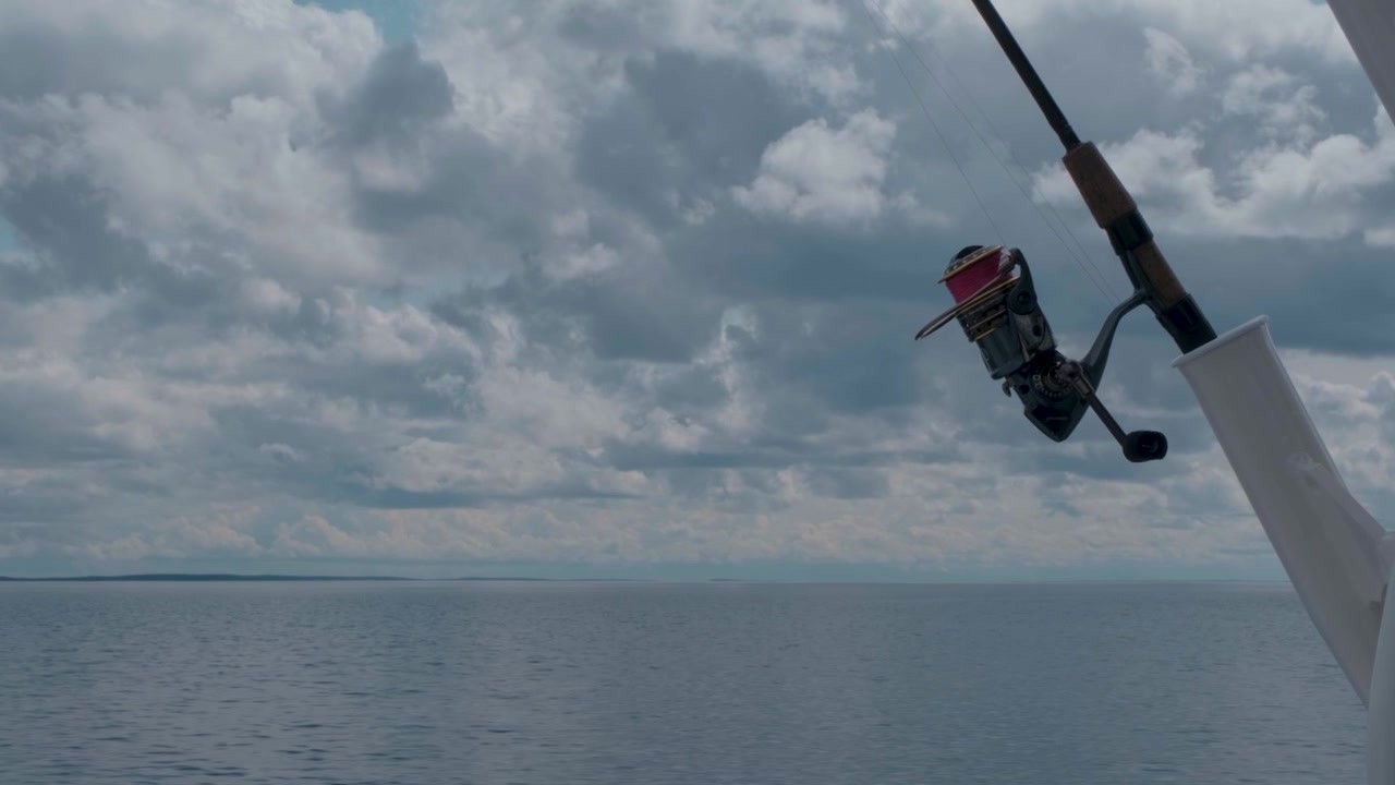 Una canna da pesca in primo piano mentre una barca naviga su un vasto lago settentrionale circondato dalla foresta