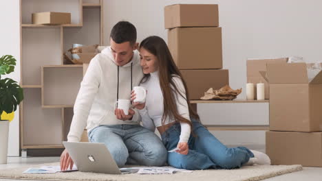 Pareja-Joven-En-Una-Casa-Nueva-Sentada-En-La-Alfombra-Con-Una-Laptop-Eligiendo-Colores-Para-La-Decoración-Mientras-Bebe-Café-1