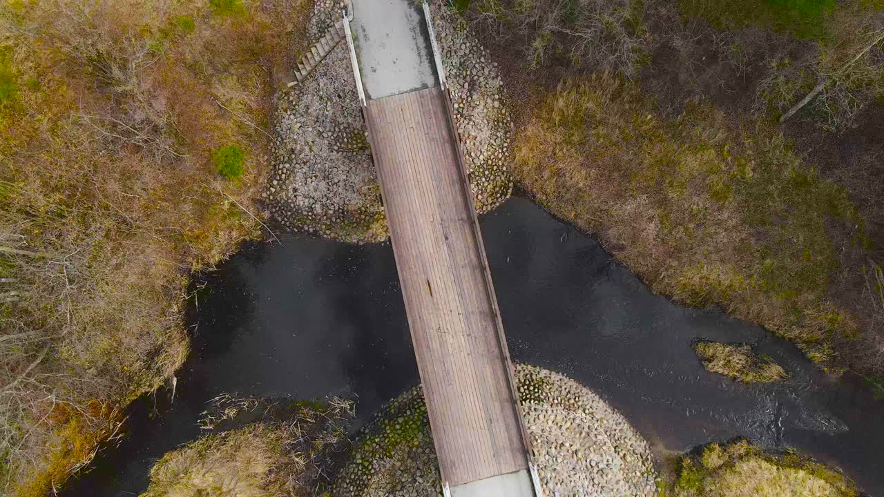 Top down view of wooden footbridge over river. High rise zoom out and rotation around bridge overpass revealing connecting pedestrian walkways. Long paths across spring scenery. Scenic route landscape
