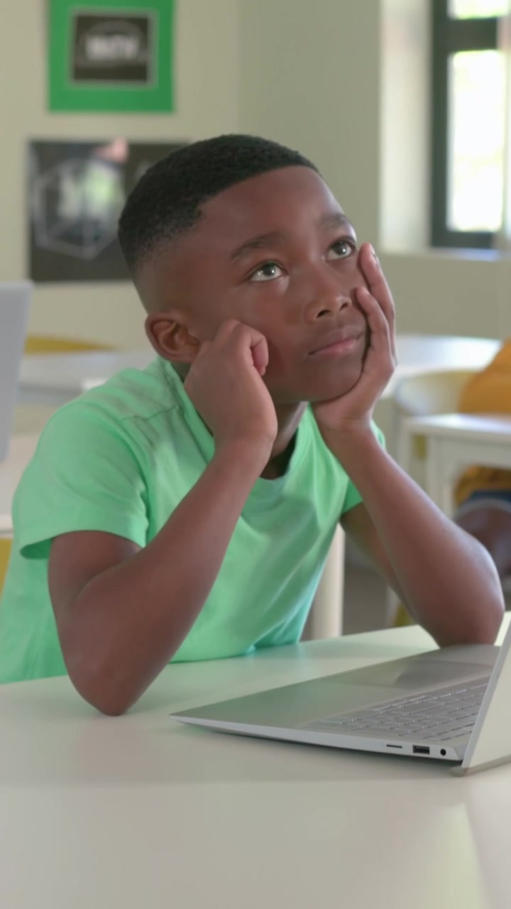 Vertical video: Typing boy in green tee doing school task, pausing, daydreaming at desk with laptop