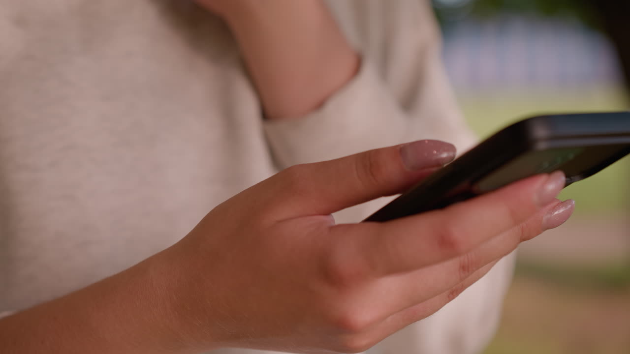 primer plano de la mano del individuo con uñas limpiamente pulidas deslizándose sobre la pantalla del teléfono inteligente, el fondo está suavemente borroso, con vegetación natural y pistas de una figura en la distancia
