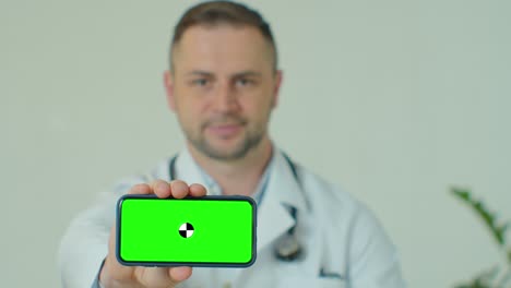médico de buen humor sosteniendo un teléfono inteligente con pantalla verde mientras está sentado en el gabinete. médico masculino demostrando teléfono con llave de croma. seguimiento de movimiento.