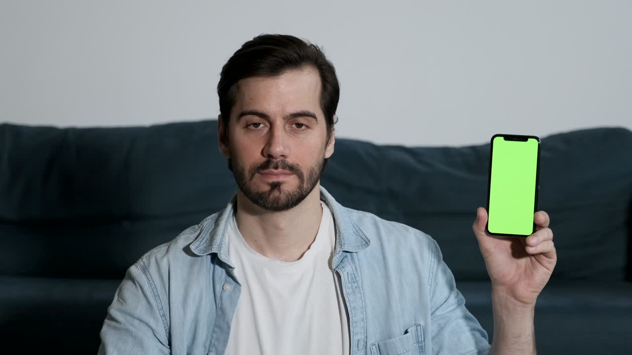 한 젊은이가 집에 있는 방에서 소파에 앉아서 카메라를 보고 새로운 응용 프로그램의 녹색 화면 통합으로 스마트폰을 보여줍니다.