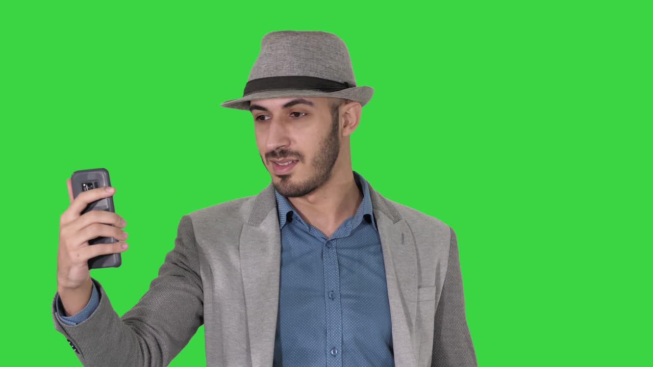 hombre atractivo con ropa casual sombrero estilista hipster grabando vlog o haciendo una llamada de video en una pantalla verde, llave de croma