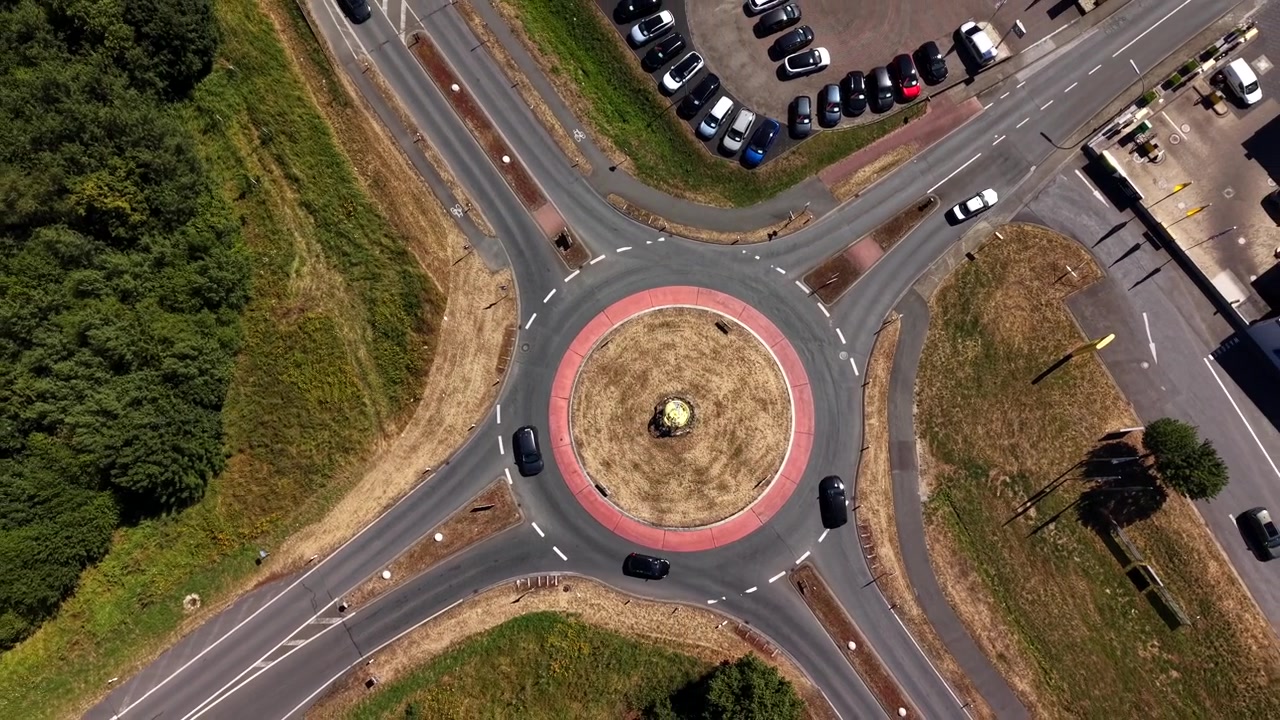 Cars circle brick-paved traffic roundabout with weathered statue centerpiece, dashed white lane markings guiding vehicles through grassy suburban USA intersection below. Aerial drone top down rising