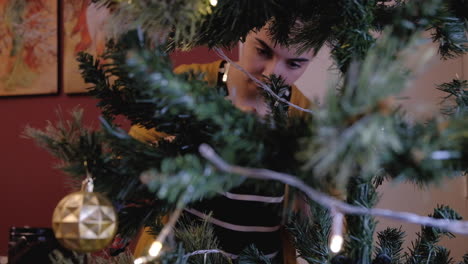 joven y hermosa mujer caucásica configurando la colocación de adornos en el árbol de navidad enmarcado entre ramas fuera de foco