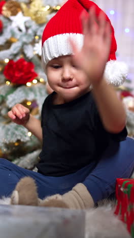 adorable kid in santa cap claps his hand by the gift boxes. baby boy sits on the floor near the christmas tree. vertical video