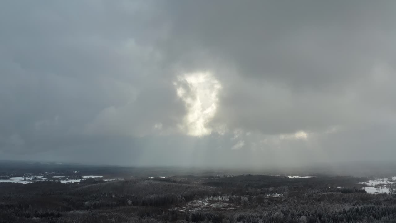 Aerial drone view of sun rays breaking through thick clouds in winter landscape. Weather in countryside with snow covered ground. Dramatic sky over the forest.