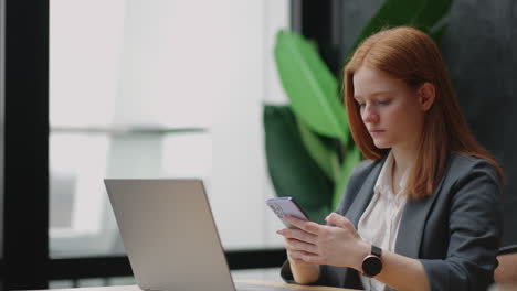 Hermosa-Joven-Mujer-De-Negocios-Usando-El-Teléfono-En-La-Oficina.-Mujer-Usando-Un-Teléfono-Inteligente-Y-Apoyada-En-Una-Ventana-Enviando-Mensajes-De-Texto-Enviando-Correos-Electrónicos-Planificando-Reuniones-Haciendo-Contactos-En-Línea-Navegando-Mensajes-En-Un-Teléfono-Móvil