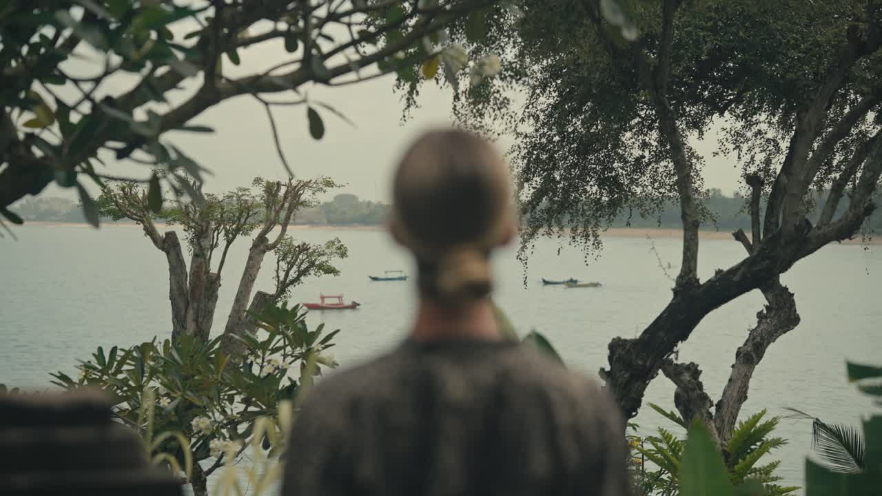 Back view of blonde woman, slightly out of focus, gazes in between trees over the ocean where traditional Balinese fishing boats float on the water