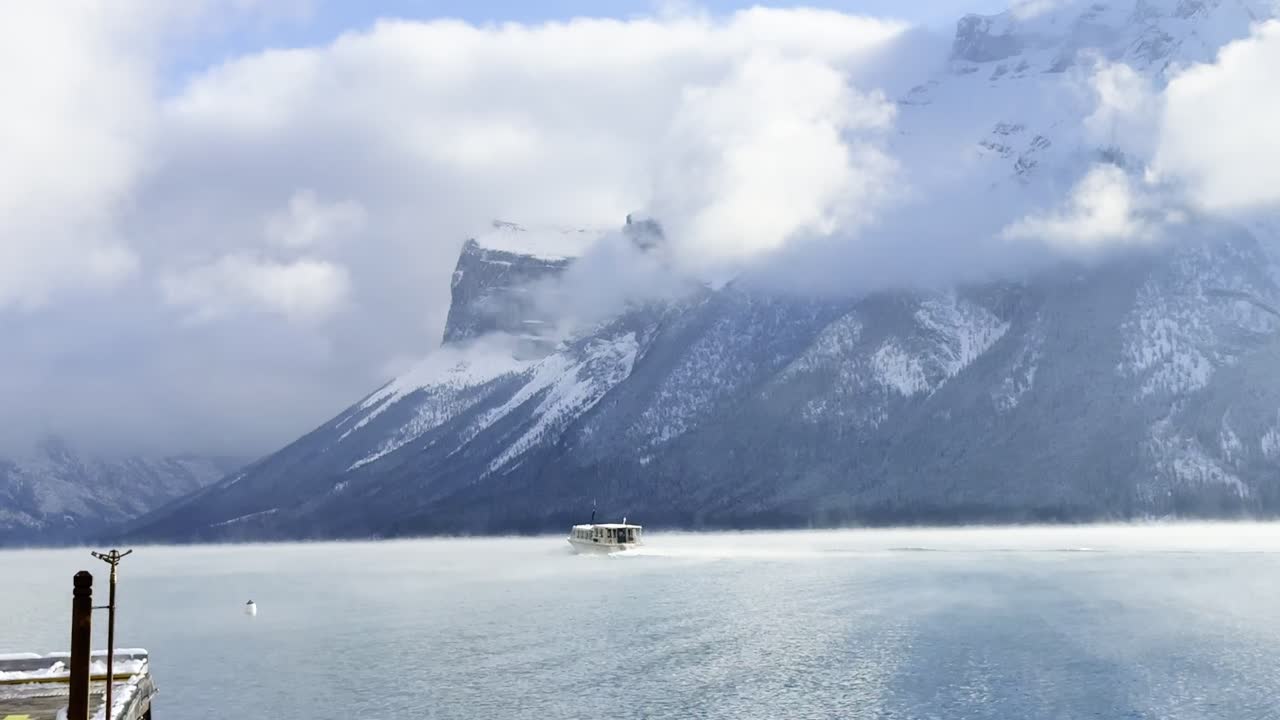 Wide view of a clear winter highway cutting through a snowy valley, lined with frosted trees and distant Rocky Mountain peaks under expansive blue sky, Alberta, Canada
