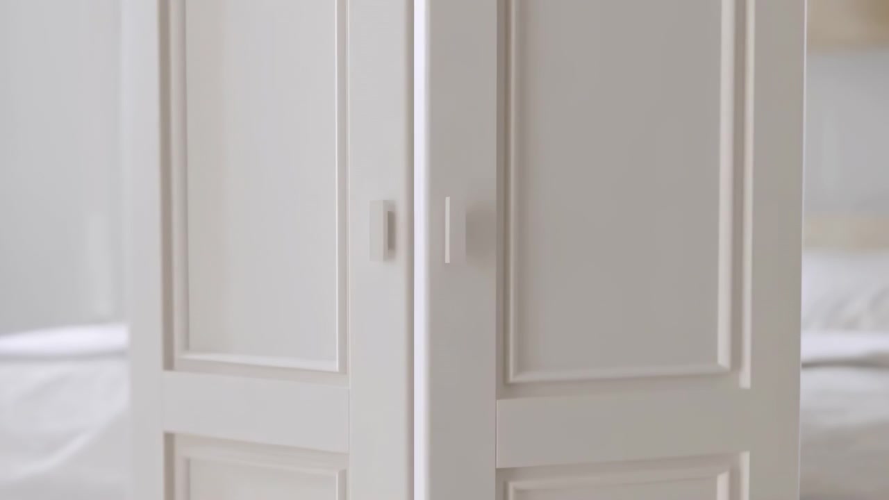 Ante di armadio bianche chiuse sono centrate in un ambiente di camera da letto calmo con luce interna soffusa, un letto sfocato ai bordi e un'atmosfera domestica pulita e minimale.