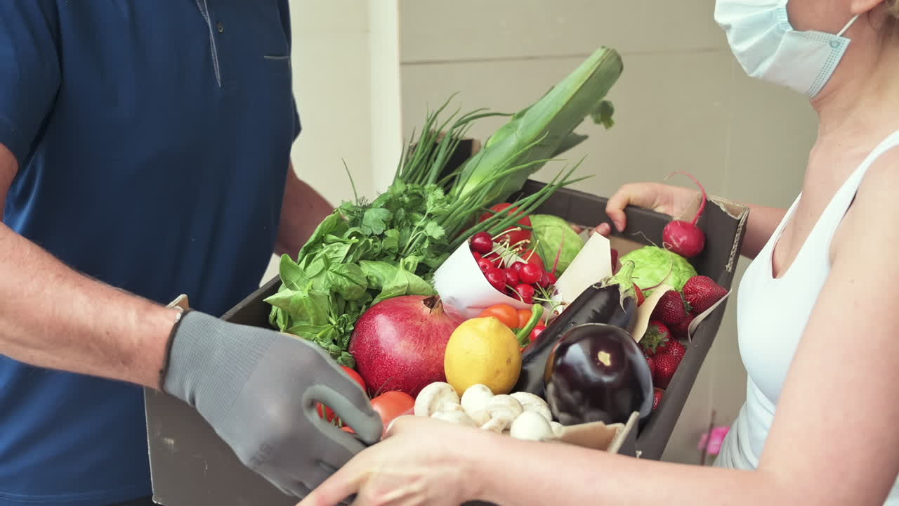 Woman receiving a box of fresh fruits and vegetables from a masked delivery man