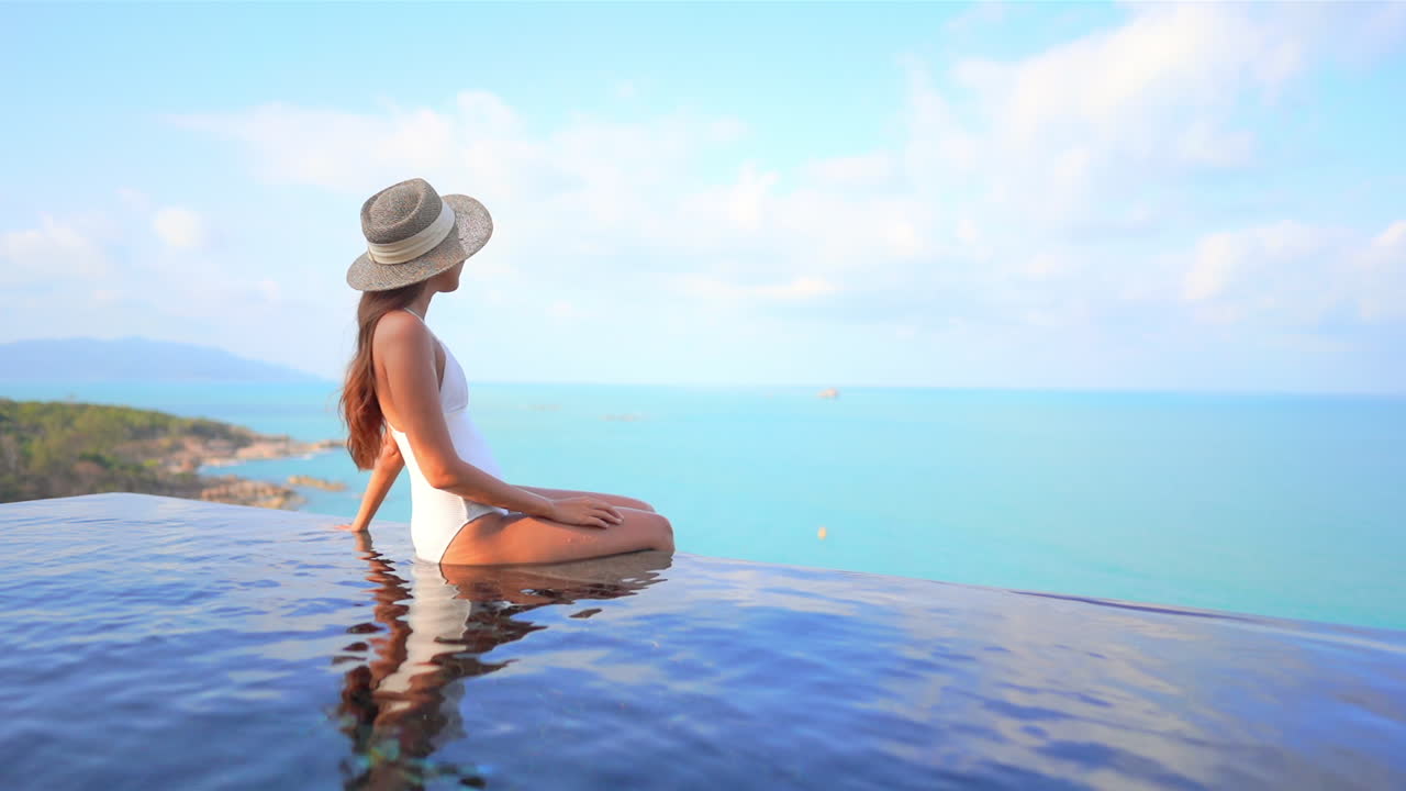Fittes asiatisches Model in Bademode sitzend am Rand des Infinity-Pools des Luxushotels auf dem Berggipfel mit herrlichem Meerespanorama, Seitenansicht