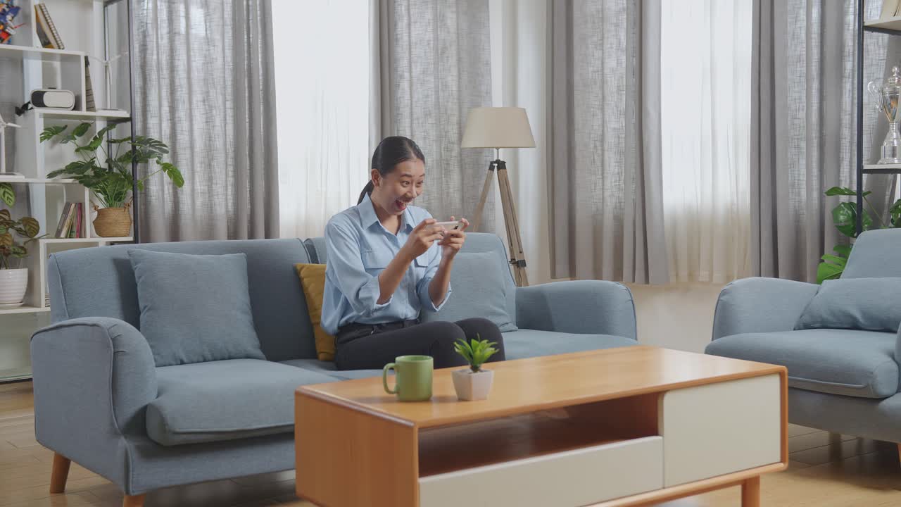 아시아 여성은 거실의 소파에 앉아 스마트폰으로 게임을 즐긴다.