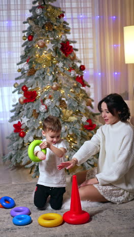 lovely baby puts a detail of toy pyramid on his head and arm. brunette woman claps happily and kid puts a toy on her wrist. mom and son near christmas tree. vertical video.