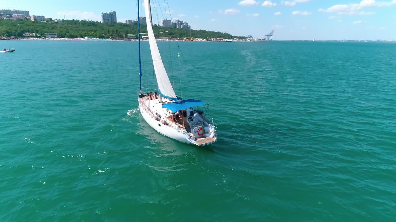 White yacht with sails floating to the shore. Beautiful sailboat with a few people on the sea in bright sunny day. Aerial view.