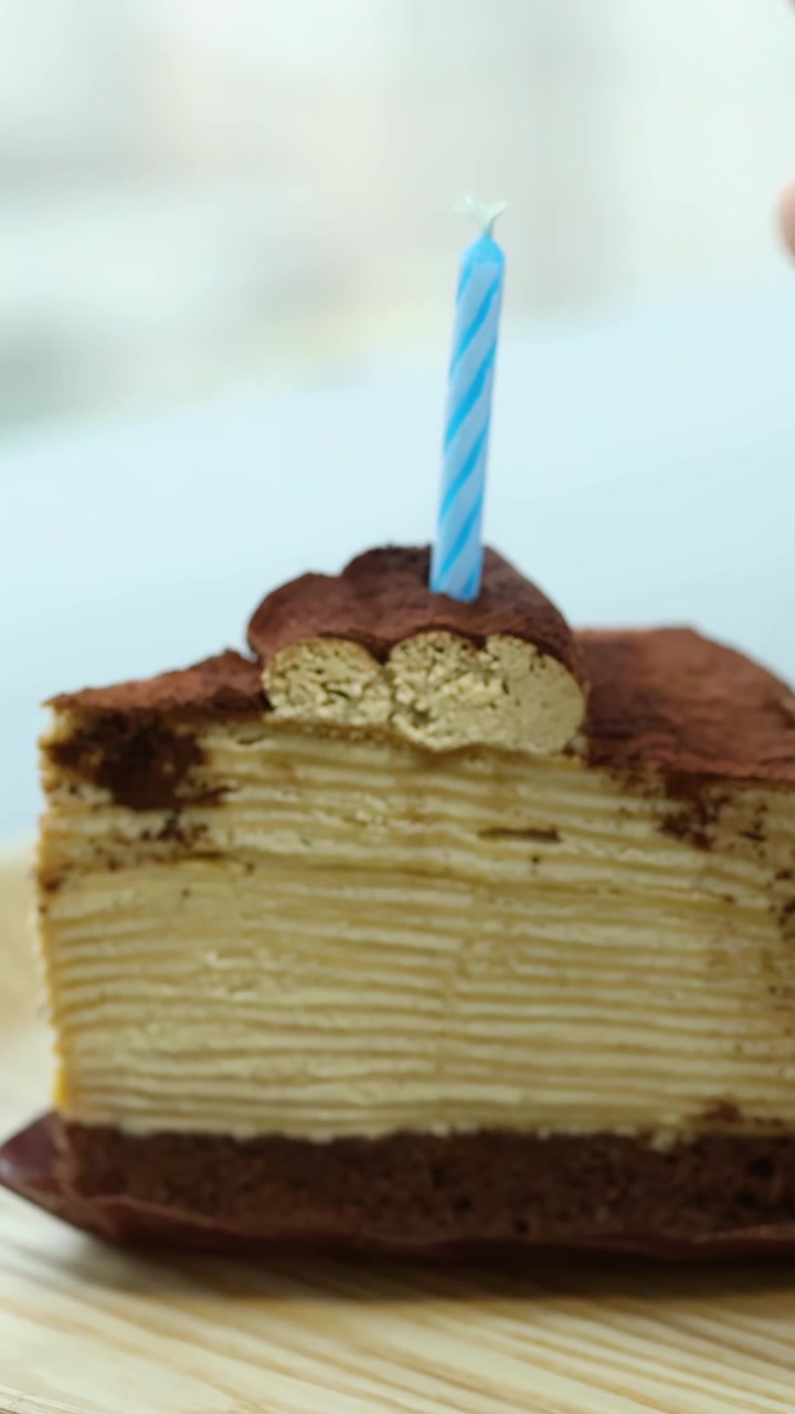 明るい自然光の下、層になったティラミスクレープケーキに青いバースデーキャンドルを置く手。