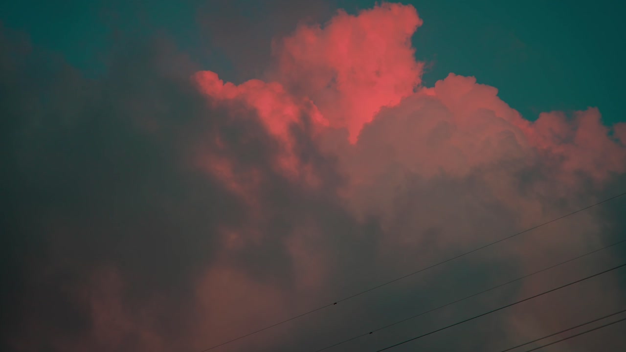 A cinematic view of thick sunset clouds glowing peach-pink against a turquoise-blue sky. Faint power lines add scale, grounding this serene and dreamlike moment