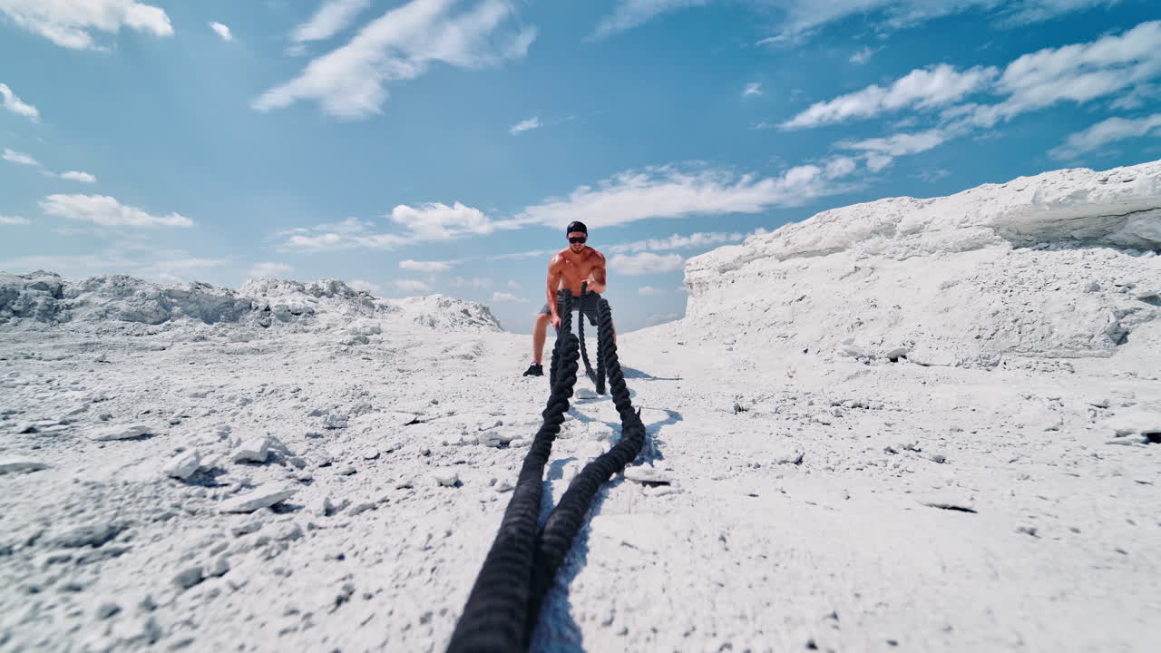 Bodybuilder trains his muscles with cable on the hill under blue sky. Shirtless sportsman wearing cap and sunglasses doing workout with battle ropes in sunny summer day outdoors.
