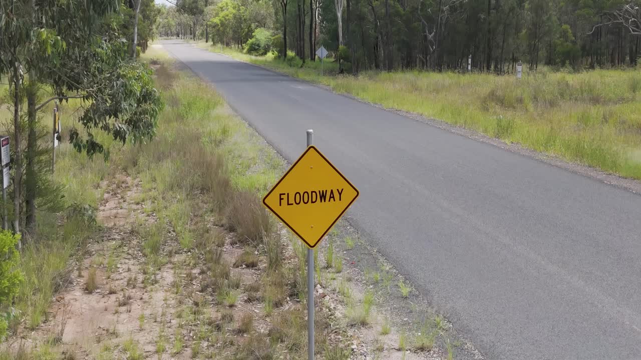 A floodway warning sign beside a rural asphalt road surrounded by grass and trees.