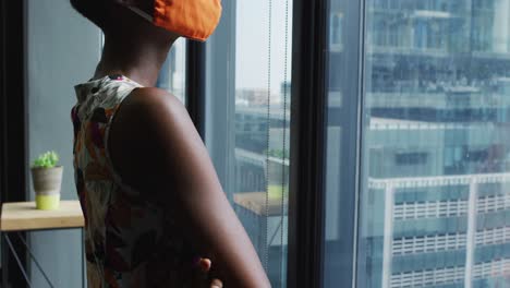 thoughtful african american woman wearing face mask looking out of window at modern office