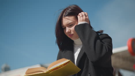 primer plano de una estudiante leyendo al aire libre, hojeando las páginas de un libro, el viento sopla suavemente su cabello, y ella ajusta su portátil. edificio de ladrillo rojo forma un fondo borroso