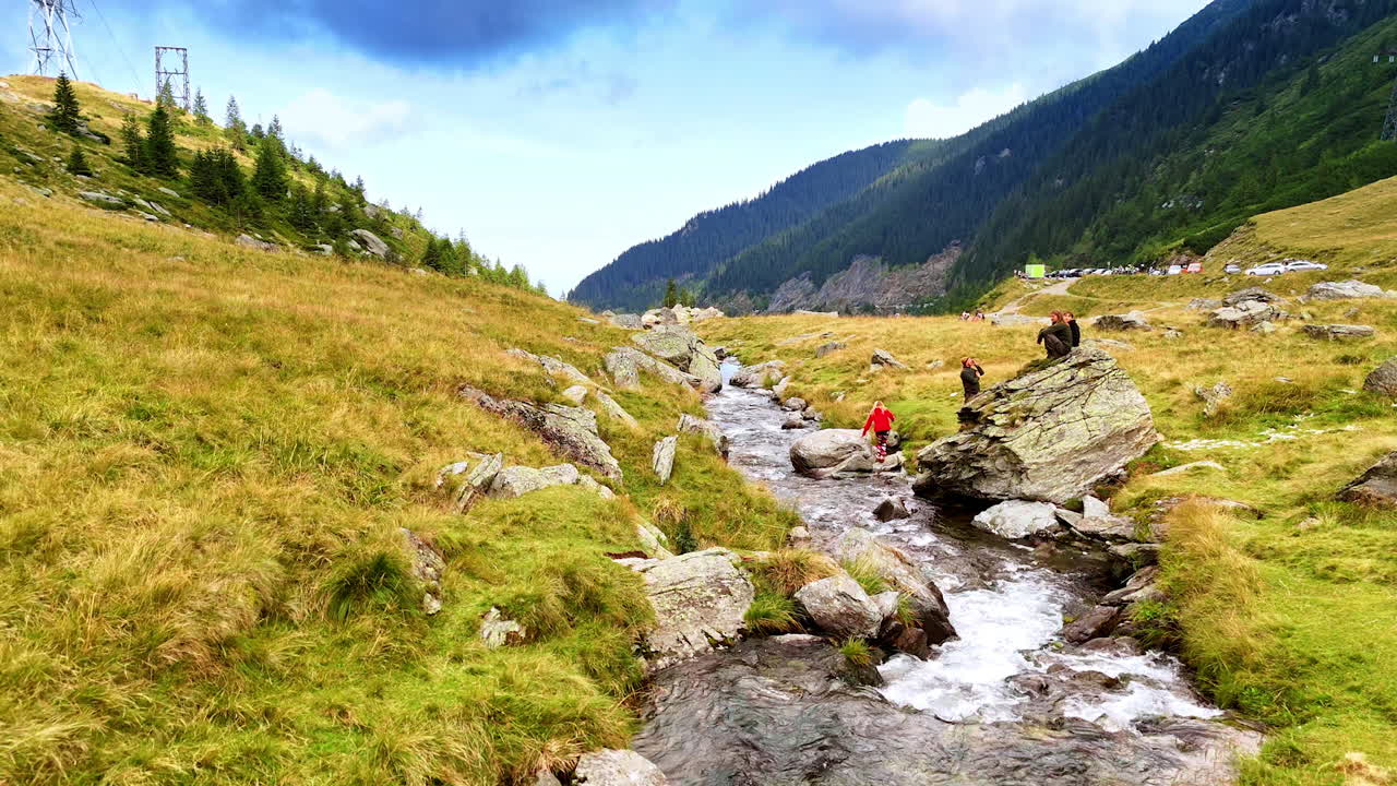 Tourists walk, rest and take pictures near the river flowing by the rocks. View on the green mountains of Romania at backdrop