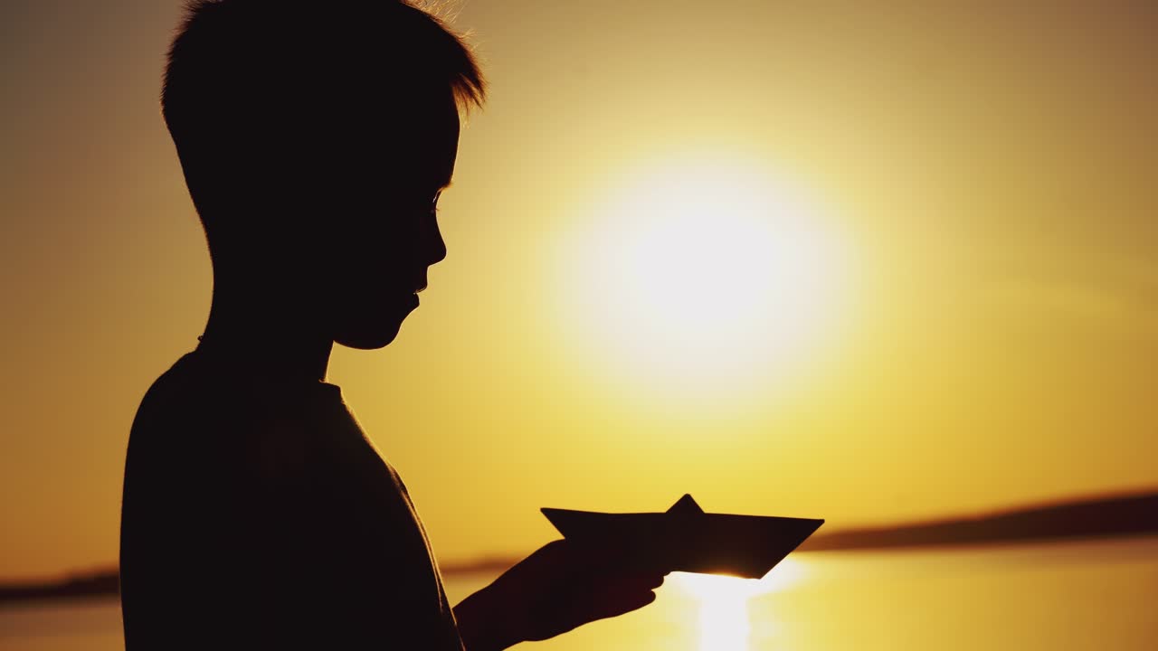 Silhouette of a little boy is holding a paper boat in his hand and looking at it on the background of sunset in the summer near the pond. Close-up