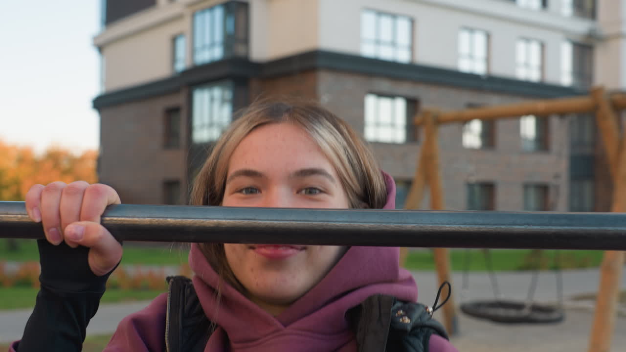 Portrait of female coach making funny face with smile while executing pull up on horizontal bar during outdoor fitness session in city playground, highlighting strength confidence and energetic mood