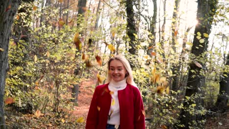 slowmotion young beautiful woman throwing leaves in the air amidst thee orange brown autumn forest woodland while wearing a red coat