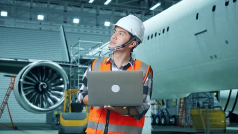 ingeniero masculino asiático con casco de seguridad mirando una computadora portátil y mirando a su alrededor mientras está de pie con aviones en el hangar